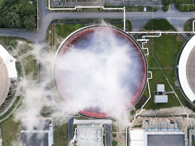 Auf der Luftaufnahme ist ein Kraftwerkskühlturm zu sehen, aus dem weißer Wasserdampf aufsteigt. Um den Kühlturm herum sind grüne Wiesen sowie Wärmerohleitungsnetze und Industriebauwerke zu erkennen.