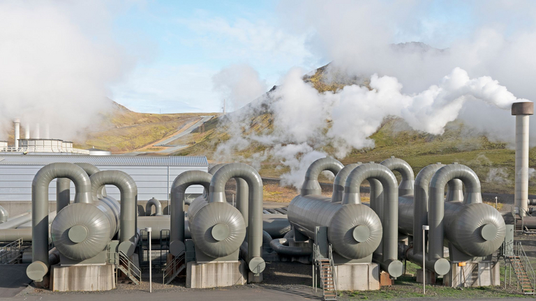Die Ansicht zeigt eine Geothermieanlage mit Lagertanks sowie Schornsteine, aus denen Wasserdampf aufsteigt. Im Hintergrund ist eine bewachsene, grüne Hügellandschaft zu sehen.