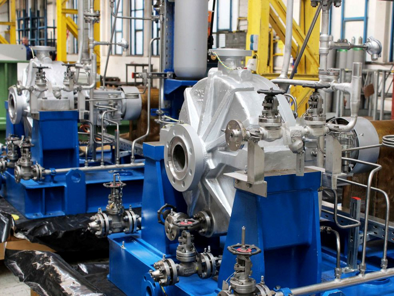 Seitlicher Blick auf zwei OH2 Prozesspumpenaggregate mit vielen verrohrungen für ein Schmierölversorgungssystem. Pumpengehäuse ist in Stahlguss, Grundrahmen ist blau lackiert und im Hintergrund sind gelbe Stahlträger der Apollo Fertigungshalle zu sehen.