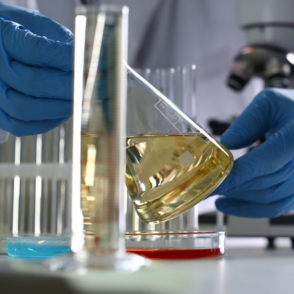 Eine Person in einem Chemielabor trägt einen weißen Kittel und blaue Schutzhandschuhe. Sie schwenkt eine chemische, gelbe Flüssigkeit in einem großen Reagenzglas.