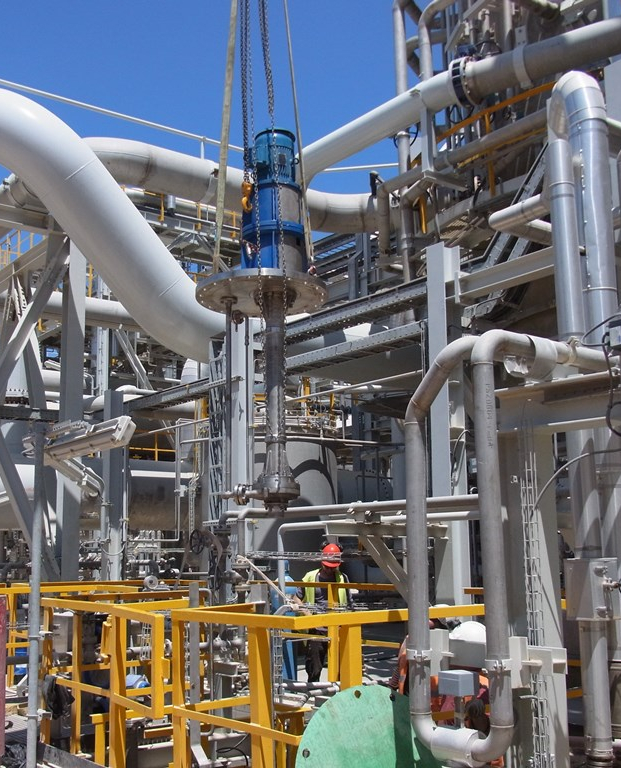 Eine vertikale einstufige KRPOV Prozesspumpe von Apollo wird in einer Chemieanlage mit dem Kran in der Anlage positioniert und montiert. Umgeben von unzähligen Verrohrungen in der Przessanlage. Im Hintergrund ist ein wolkenloser, blauer Himmel zu sehen.