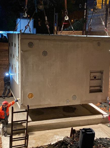 Zu sehen ist eine Baustelle bei Nacht, auf der ein Wassercontainer mithilfe eines Krans von einer Druckerhöhungslage auf ein Betonfundament gesetzt wird. Zwei Arbeiter in leuchtender Arbeitskleidung helfen bei der genauen Positionierung.