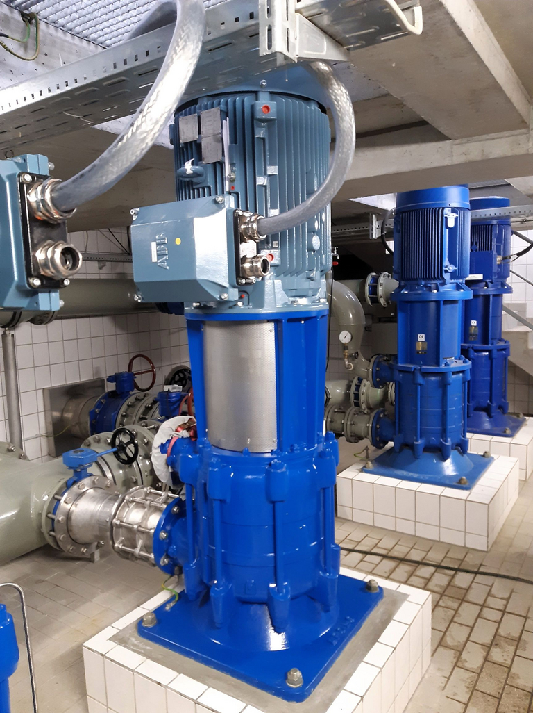 Seitlicher Blick in eine Trinkwasseranlage mit Anschlüssen, Wasserleitungen und nebeneinander stehende trockenaufgestellte Apollo mehrstufige vertikale GLVB Kreiselpumpen. Fußboden ist gefliest in einer Kellerähnlichen Umgebung.