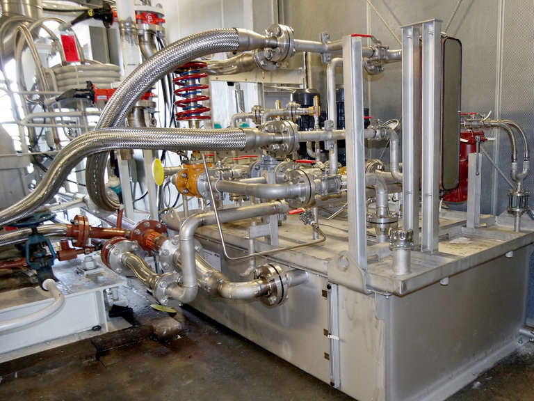 Here you can see an Apollo lubricating oil system in use at a power plant. The image shows a compact module with an oil tank, double filters, and cooling unit, which is installed and connected with numerous hoses and pipes and wired to an industrial plant in the background.